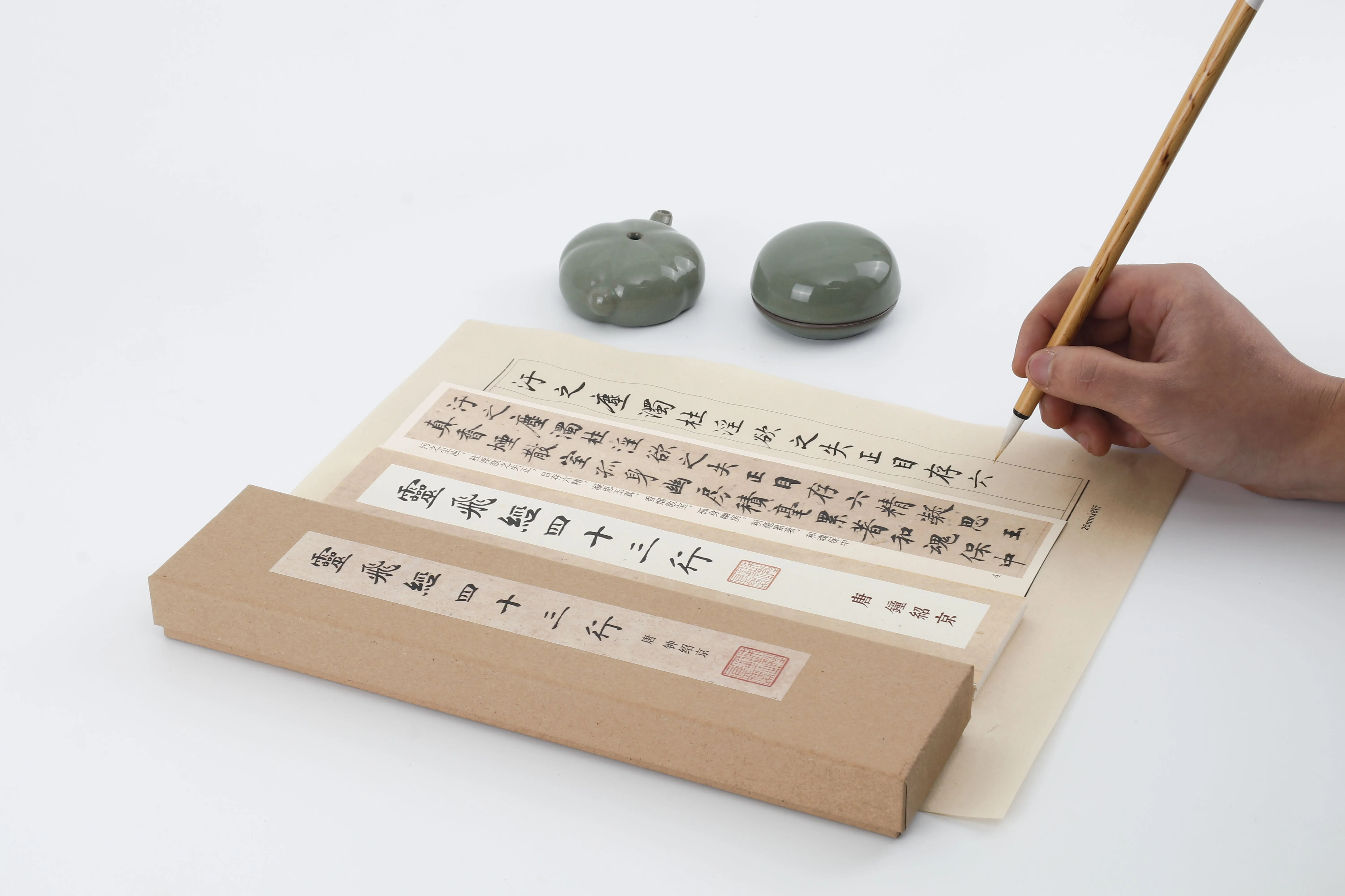 Hands writing on folded xuan paper beside a celadon water pot and a reproduction of the Lingfei Jing album
